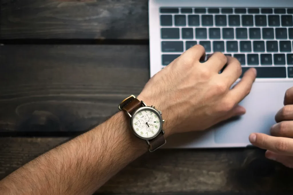 Mão masculina com relógio de pulseira de couro digitando em teclado de laptop prateado sobre uma mesa de madeira.