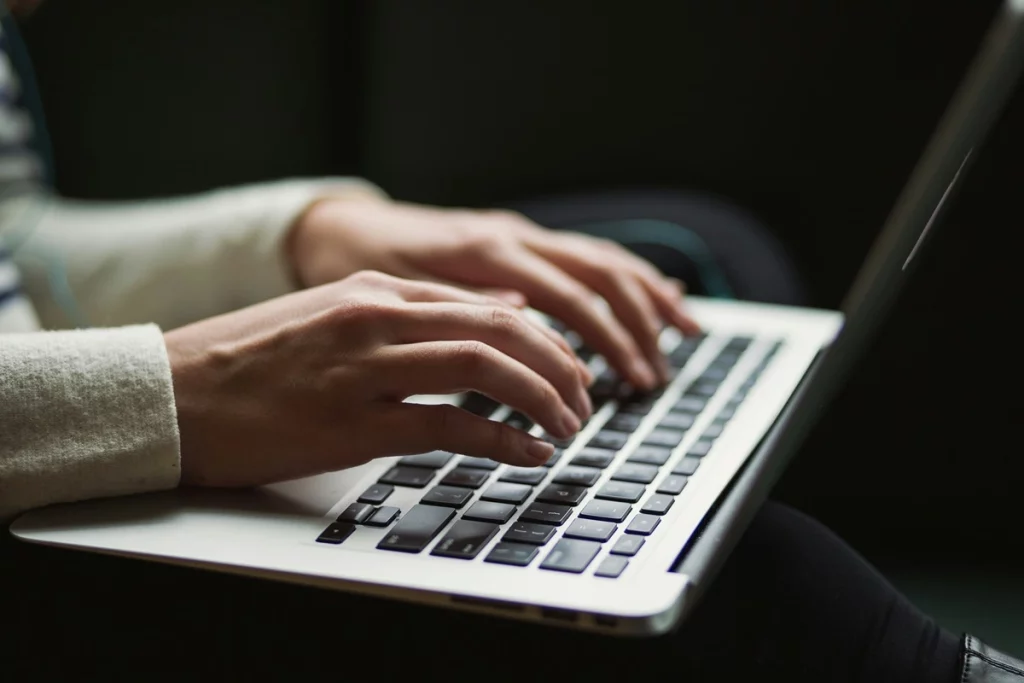 Mãos femininas digitando em teclado de laptop prata, representando trabalho, estudo ou atividades digitais