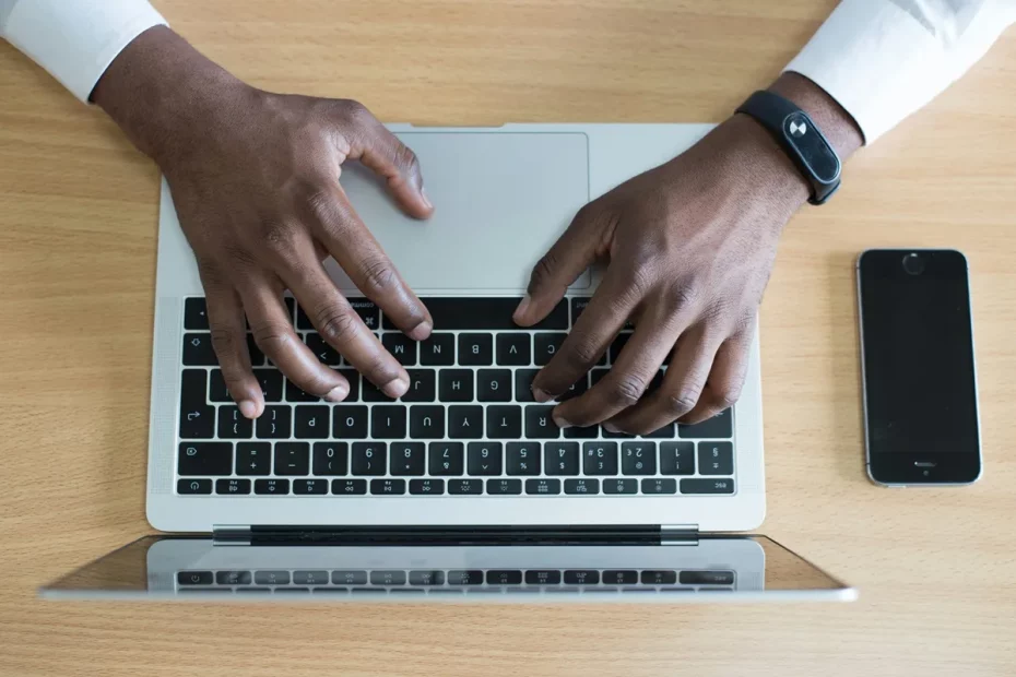 Mãos masculinas digitando em um laptop prateado sobre uma mesa de madeira, com um smartphone preto ao lado.