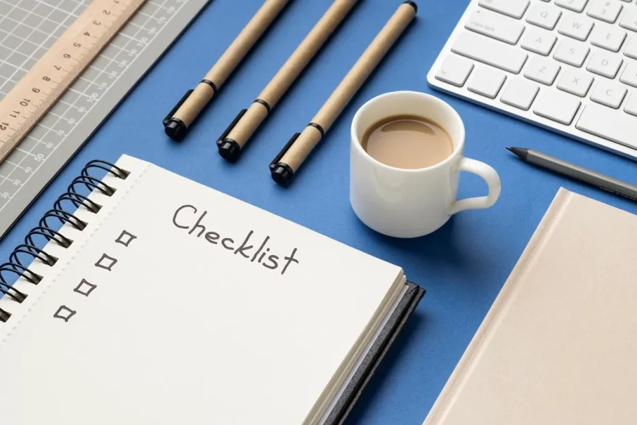 Mesa azul com um caderno de checklist aberto, três canetas de papelão ecológico, régua, teclado branco, xícara de café com leite e itens de papelaria organizados.