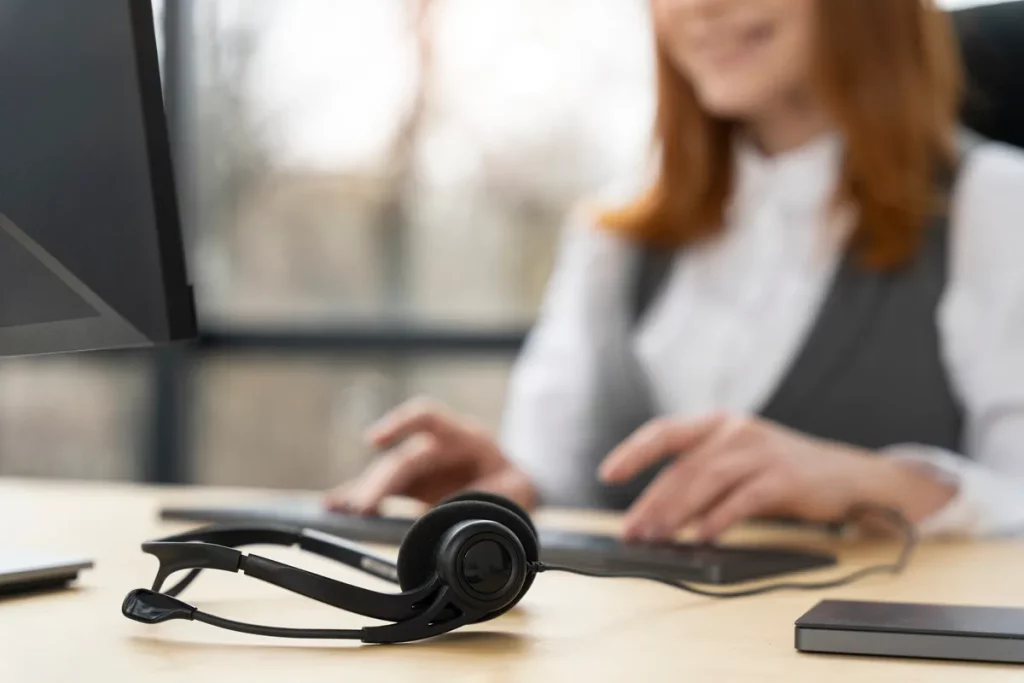 Headset preto sobre uma mesa de escritório com uma profissional desfocada ao fundo digitando no computador