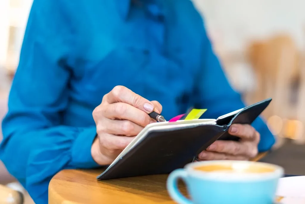 Pessoa vestindo camisa azul escrevendo em uma agenda com marcadores coloridos, ao lado de uma xícara de café em uma mesa de madeira.