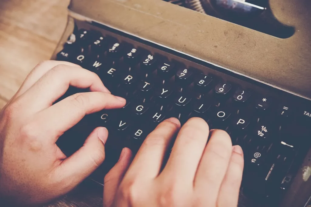 Mãos digitando em uma máquina de escrever antiga com teclas pretas e detalhes metálicos, sobre uma mesa de madeira.