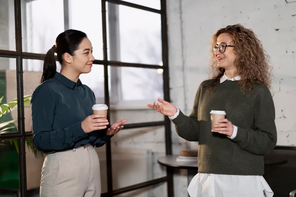 Duas mulheres conversando amigavelmente enquanto seguram copos de café em um ambiente de trabalho com paredes brancas e janelas amplas.