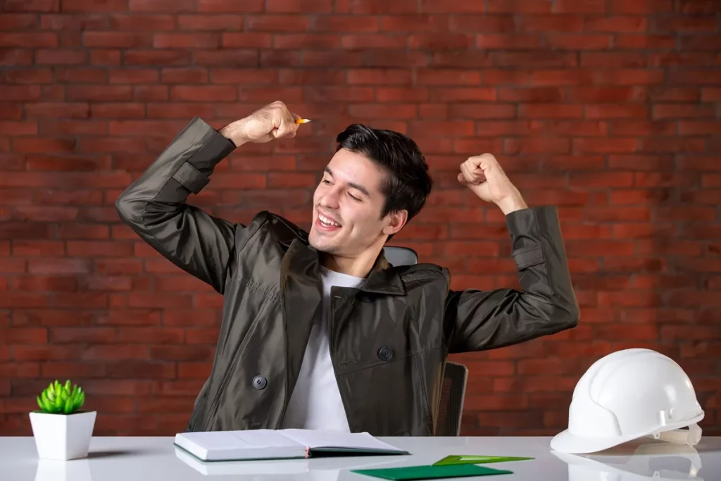 Homem jovem, vestindo jaqueta escura, comemorando com os braços levantados e sorriso, sentado em uma mesa de trabalho com caderno aberto e capacete branco sobre a mesa.