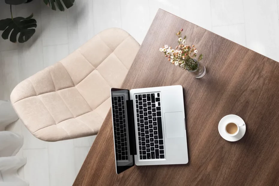 Vista aérea de uma mesa de madeira com laptop aberto, xícara de café e pequeno vaso com flores brancas, ao lado de uma cadeira estofada bege.