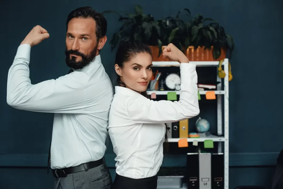 Homem e mulher de costas um para o outro, flexionando os braços em sinal de força e confiança, vestindo camisas sociais brancas, em um escritório com prateleiras ao fundo.