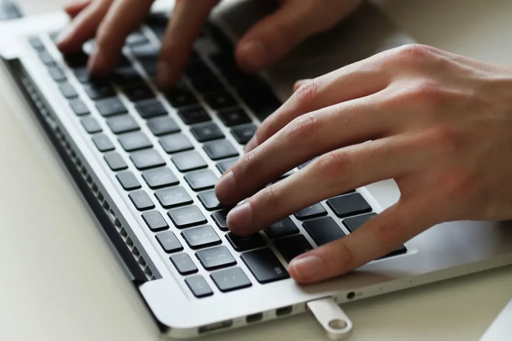 Mãos masculinas digitando em um notebook prateado com teclas pretas, transmitindo foco e precisão.