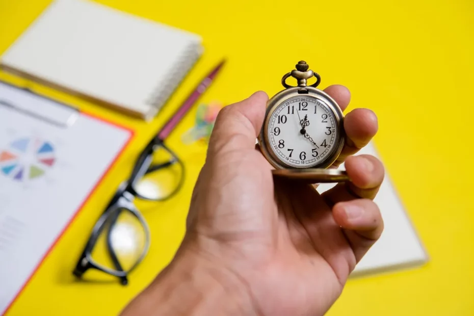 Mão segurando um relógio de bolso vintage em fundo amarelo, com itens de escritório ao redor, como óculos, caderno, caneta e gráfico.