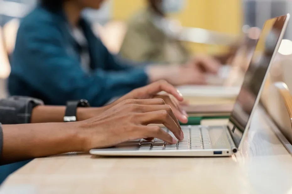 Mãos digitando em teclado de laptop em uma sala de aula com várias pessoas ao fundo utilizando computadores.