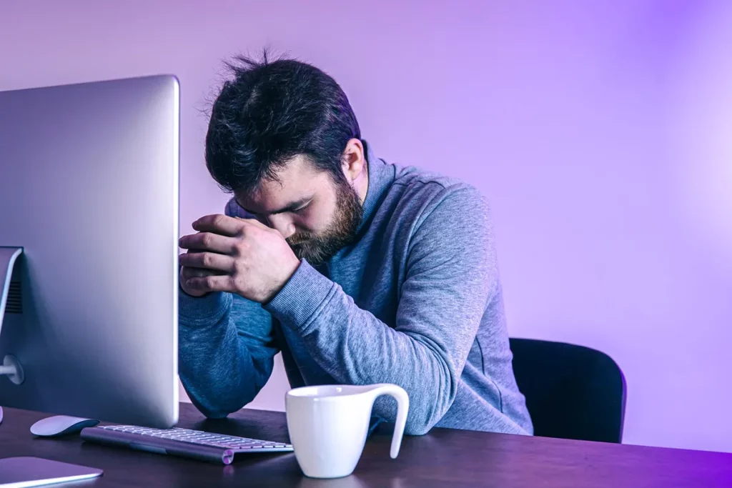 Homem com expressão de estresse e frustração apoiado na mesa em frente ao computador, com as mãos unidas e a cabeça baixa.