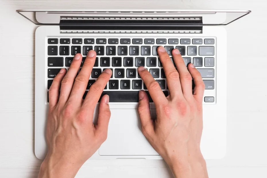 Mãos digitando em um teclado de notebook visto de cima, com um fundo branco. O teclado apresenta um layout padrão QWERTY com teclas pretas e letras brancas.