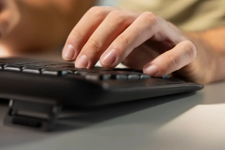 Mão masculina digitando em um teclado preto ergonômico com suporte ajustável, em um ambiente de trabalho iluminado.