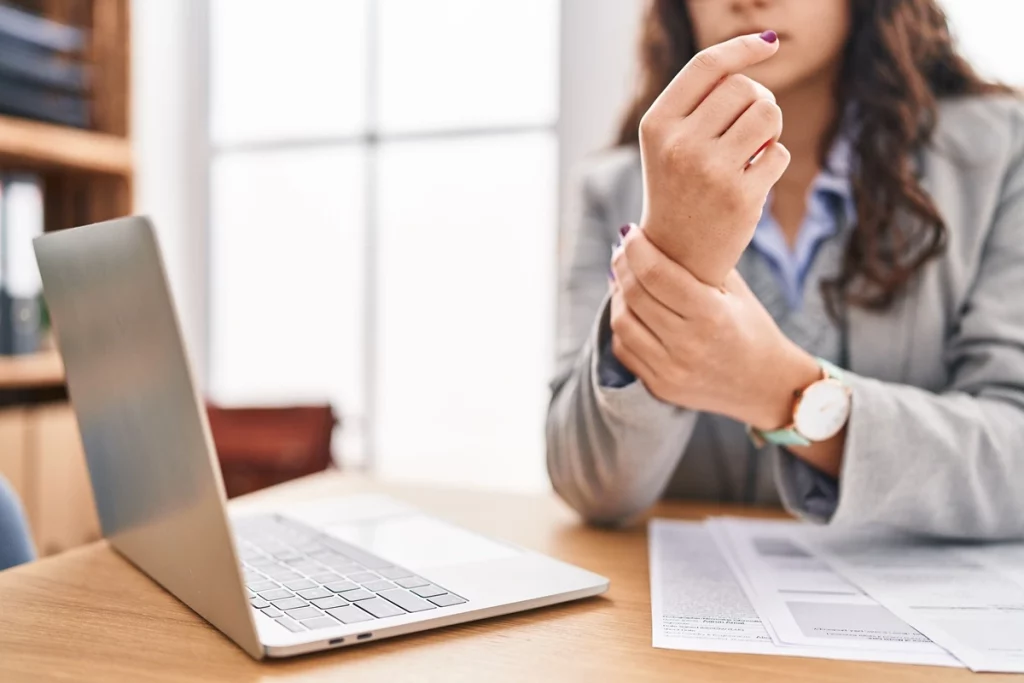 Mulher em escritório segurando o pulso com expressão de desconforto, possivelmente devido ao esforço repetitivo no trabalho.