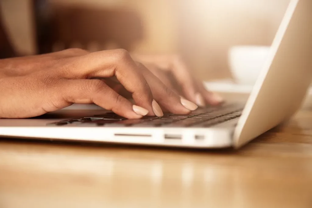 Mãos femininas digitando em um notebook branco sobre uma mesa de madeira, com iluminação suave e ambiente aconchegante ao fundo.