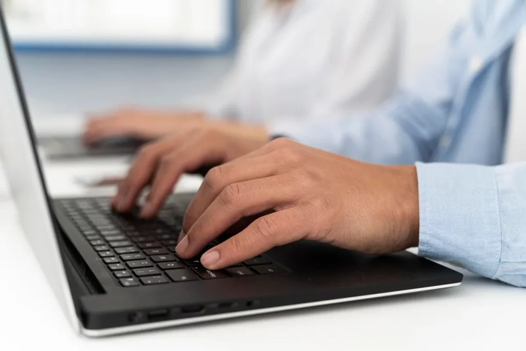 Mãos masculinas digitando em um notebook preto, com outra pessoa ao fundo também trabalhando em um laptop, representando um ambiente profissional e colaborativo.