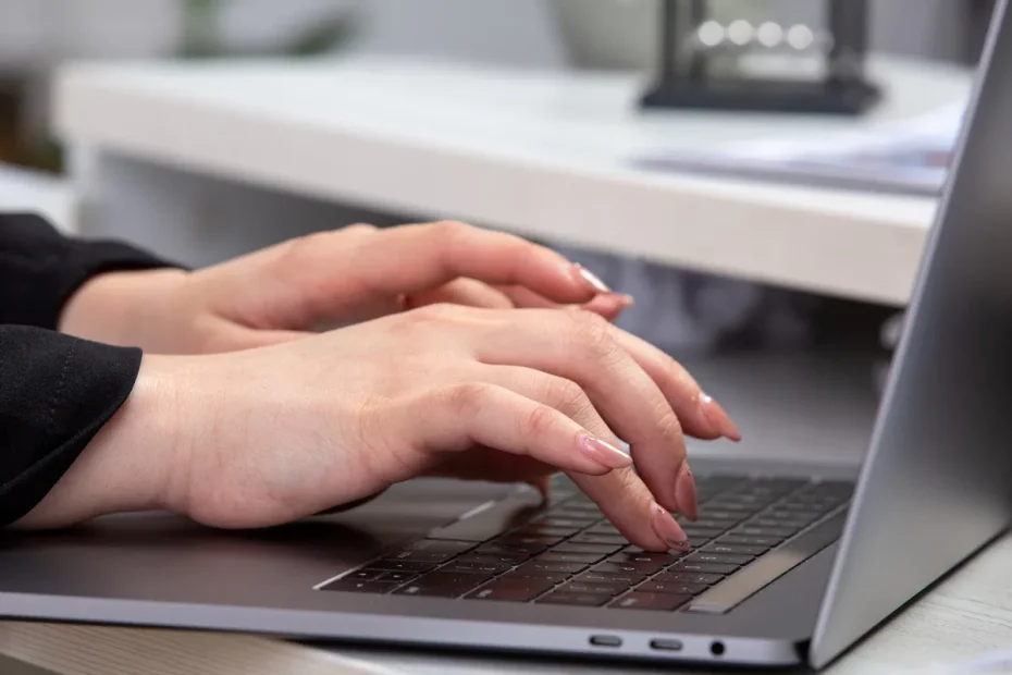 Mãos femininas digitando em um notebook prateado sobre uma mesa branca, com unhas bem cuidadas e arredondadas, sugerindo um ambiente de trabalho organizado e profissional.